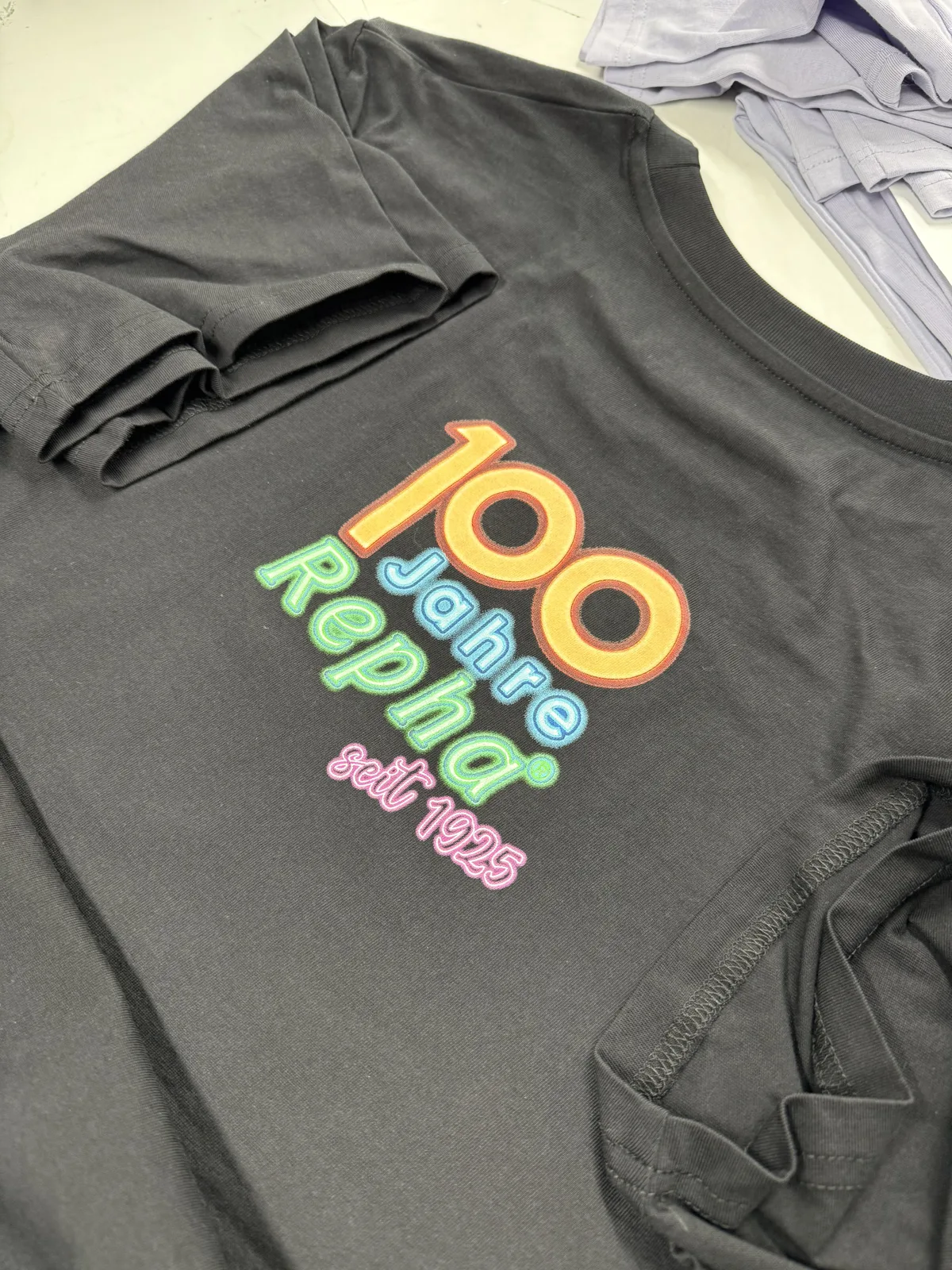 Dunkelgraues Jubiläumsshirt mit mehrfarbigem Logo zum 100-jährigen Firmenjubiläum im Siebdruck bei TIME/LESS Prints.